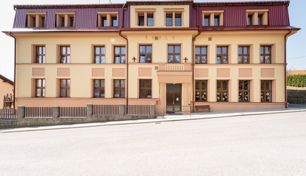 Béžovo-červená třípatrová budova s mnoha velkými okny a nápisem ŠKOLA nad vchodem, stojící za slunečného dne vedle tiché, prázdné ulice.