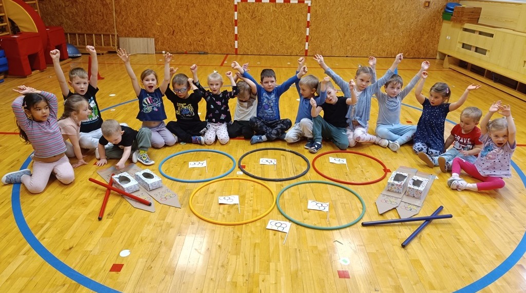 Skupina malých dětí sedí na podlaze tělocvičny v půlkruhu a zvedá ruce. Před nimi leží barevné obruče, čísla na papíře a předměty z kartonu uspořádané do herní nebo činnostní sestavy.
