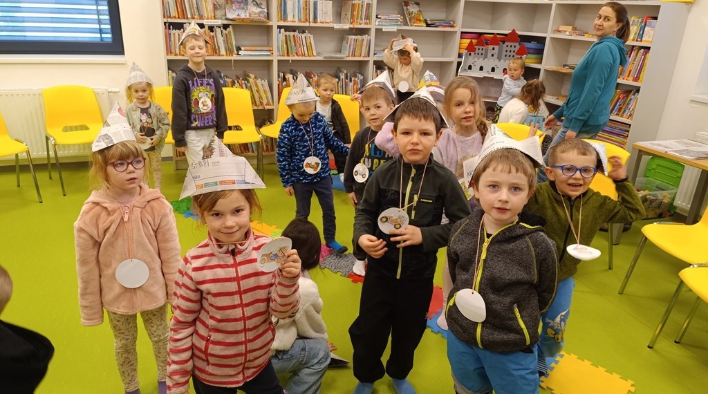 Skupina malých dětí s ručně vyrobenými papírovými čepicemi a kulatými papírovými odznaky stojí a usmívá se v jasně osvětlené místnosti knihovny s regály s knihami a žlutými židlemi v pozadí. Dospělý stojí opodál a pozoruje je.