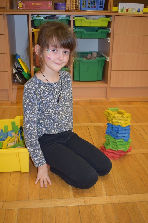 Mladá dívka s pomalovaným obličejem sedí na dřevěné podlaze ve třídě a usmívá se vedle hromady barevných dřevěných kostek a žlutého koše plného dalších kostek. V pozadí jsou police se zelenými koši.