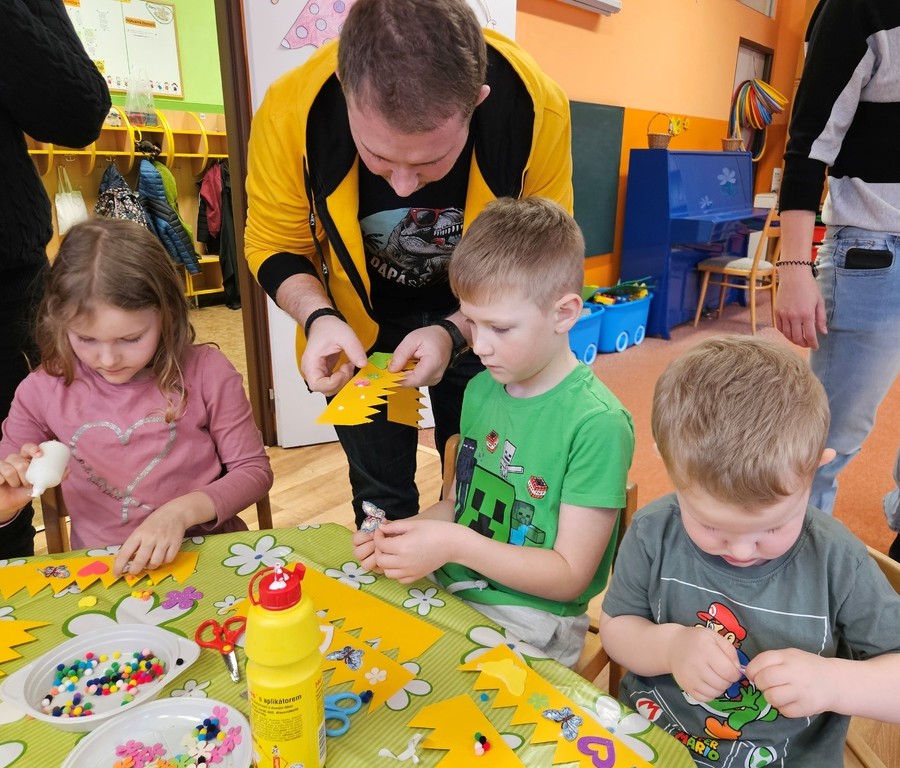 Dospělá osoba pomáhá třem malým dětem s řemeslnou činností u stolu, při níž se používá barevný papír, lepidlo, korálky a další dekorace ve třídě.