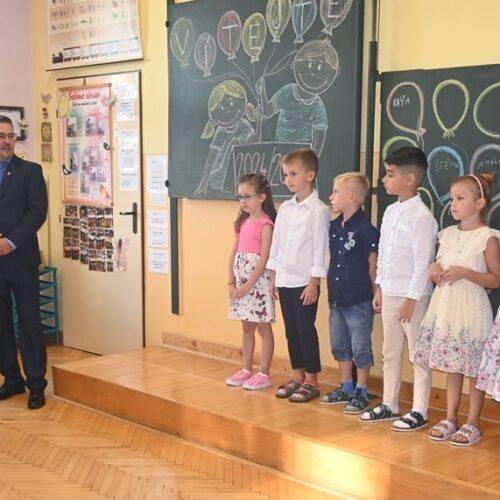 Skupina malých dětí stojí v řadě na pódiu ve třídě a poblíž stojí dva dospělí. Za nimi je tabule vyzdobená křídou s kresbami balónků a dětí. Třída je světlá a barevná.