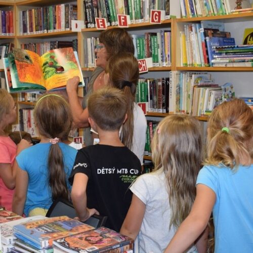 Žena čte obrázkovou knížku skupině malých dětí v knihovně obklopené regály plnými knih. Děti stojí a pozorně se dívají na čtenářku.