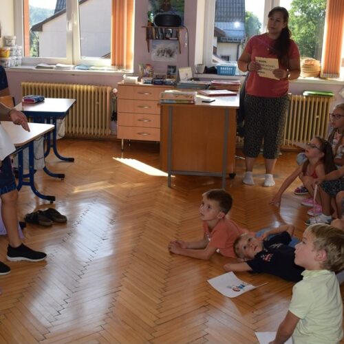 Dvě dívky stojí v čele jasně osvětlené třídy a přednášejí skupině sedících dětí. Dospělý stojí opodál a pozoruje je. Děti vypadají zaujatě a drží v rukou papíry.